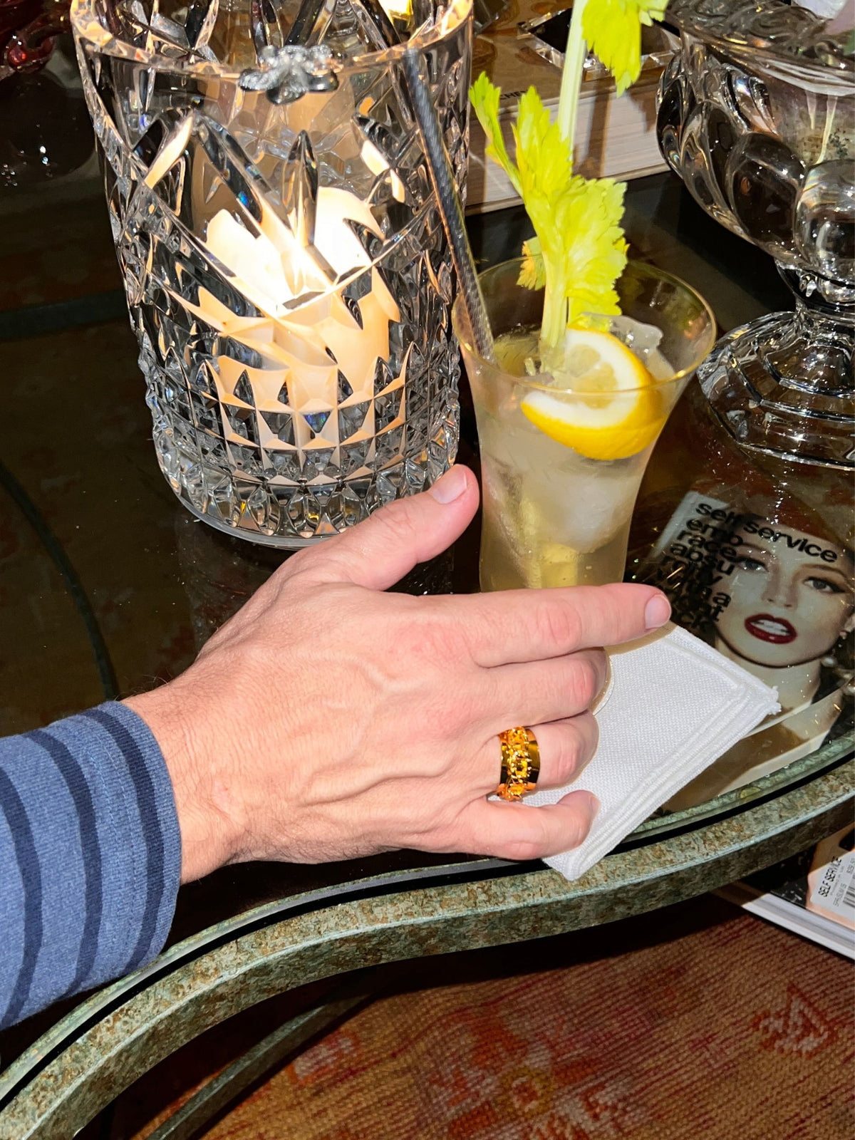 A person wearing JAREDJAMIN Jewelry&#39;s Gold Charlie Chain Mens Ring in size 7, with a striped sleeve, holds a drink with lemon and celery. The drink is on a napkin beside a lit candle in a crystal holder and beneath the glass table is a book featuring a woman&#39;s face.