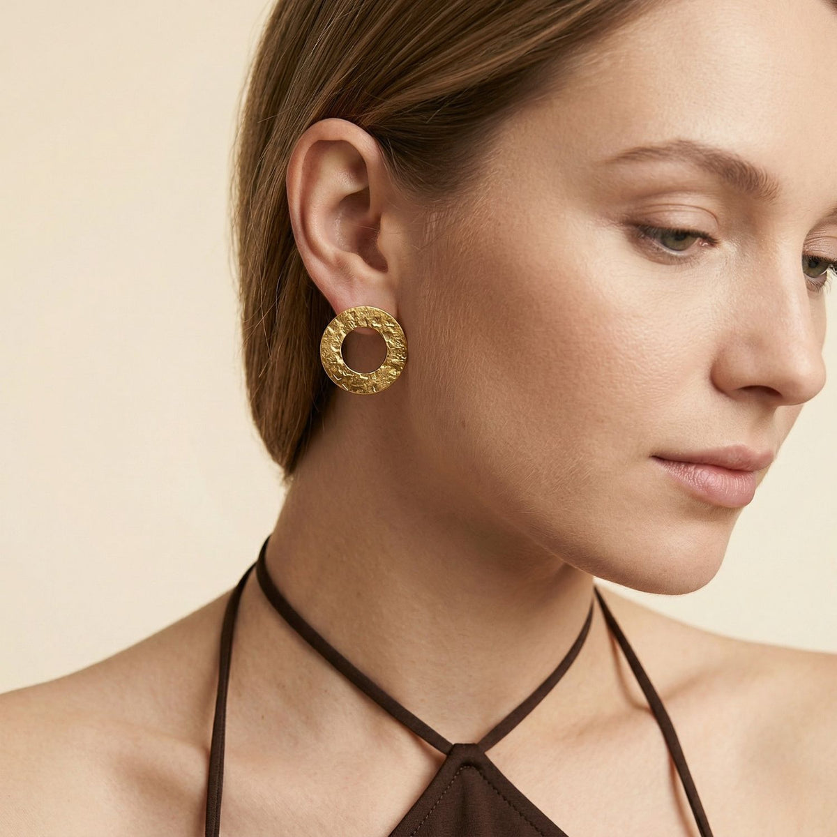 oro Colorado pendientes de JAREDJAMIN presentan a signature brushed finish, crafted from sustainable materials in small batches in West Hollywood. Un woman models them en un brown halter top against a light beige backdrop con minimal makeup.