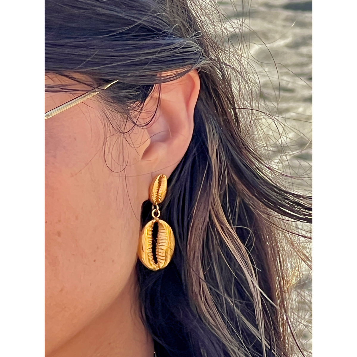 gros plan of a person wearing JAREDJAMIN Bijoux en Ligne’s or Cowrie pendante boucles d'oreilles, 18K or plated et shaped like cowrie shells. Their highlighted dark hair partly covers the ear, avec a blurred background resembling a sandy summer beach.