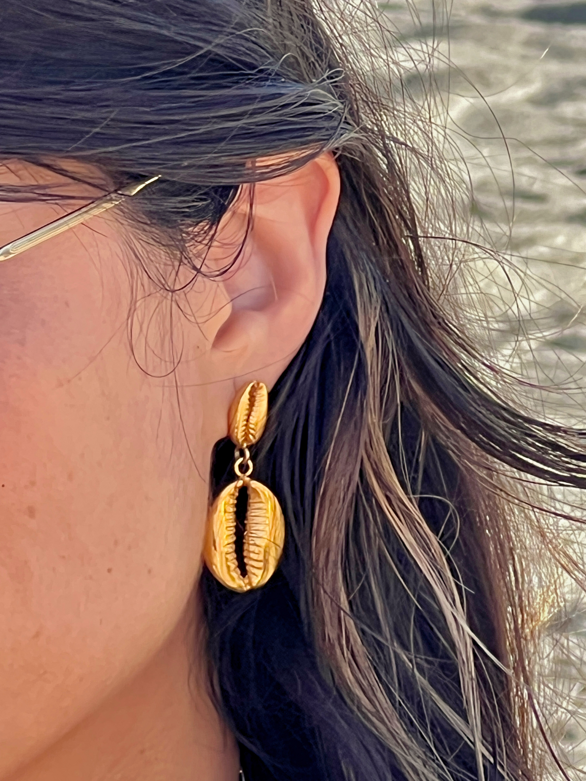 Close-up of a person wearing JAREDJAMIN Jewelry Online&#39;s Gold Cowrie Drop Earrings, elegantly crafted with 18K gold plating. Two shells are featured, a smaller one atop a larger. The person has dark hair with lighter streaks and wears glasses against a subtly textured background.