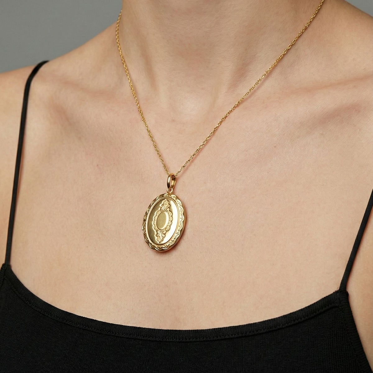 Wearing the JAREDJAMIN Jewelry Online Gold Edwardian Necklace Pendant Locket, a person in a black spaghetti strap top poses against a gray background, focusing on the neck, collarbone, and upper chest.