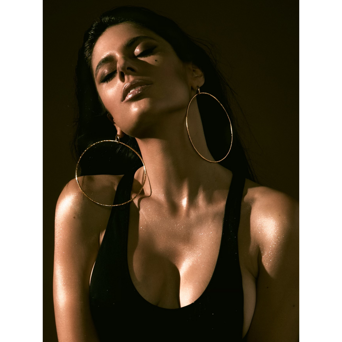 Un woman avec long dark hair et glowing skin poses avec eyes closed, wearing a black tank top et Ellen Large texturé créole Post boucles d'oreilles par JAREDJAMIN Bijoux en Ligne. Dramatic, warm light highlights her face et shoulders against a dark background.