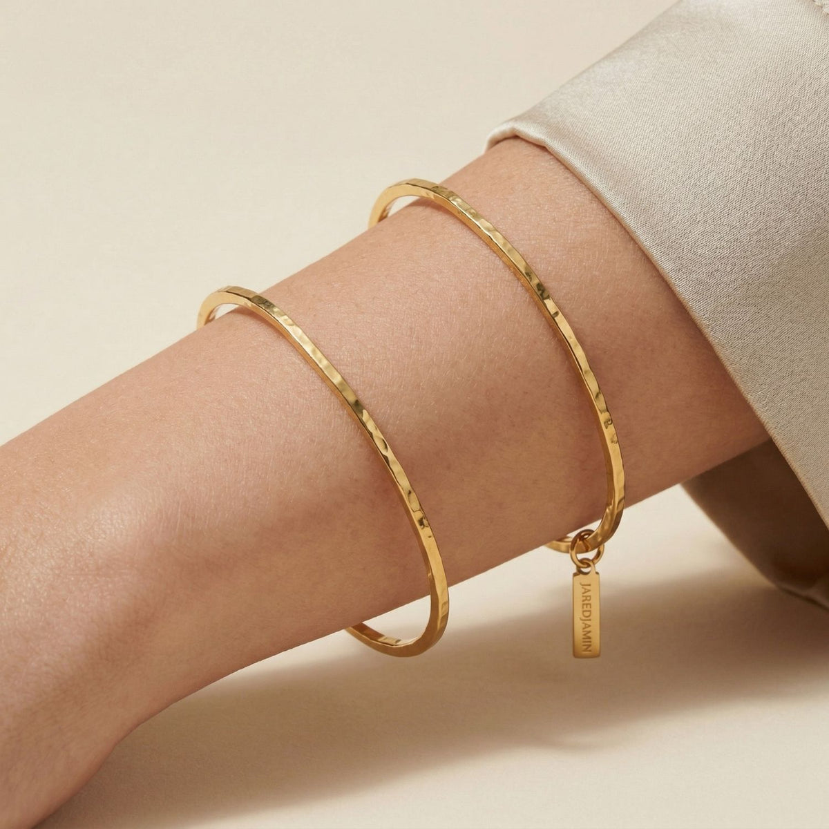 A wrist punctuates the frame, adorned with two gold bangles—one, our hand-assembled Gold Halo Forge Textured Cuff from West Hollywood, refracts light with hammered intricacy. Crafted in small batches, each piece transitions effortlessly from suiting to slip dresses.