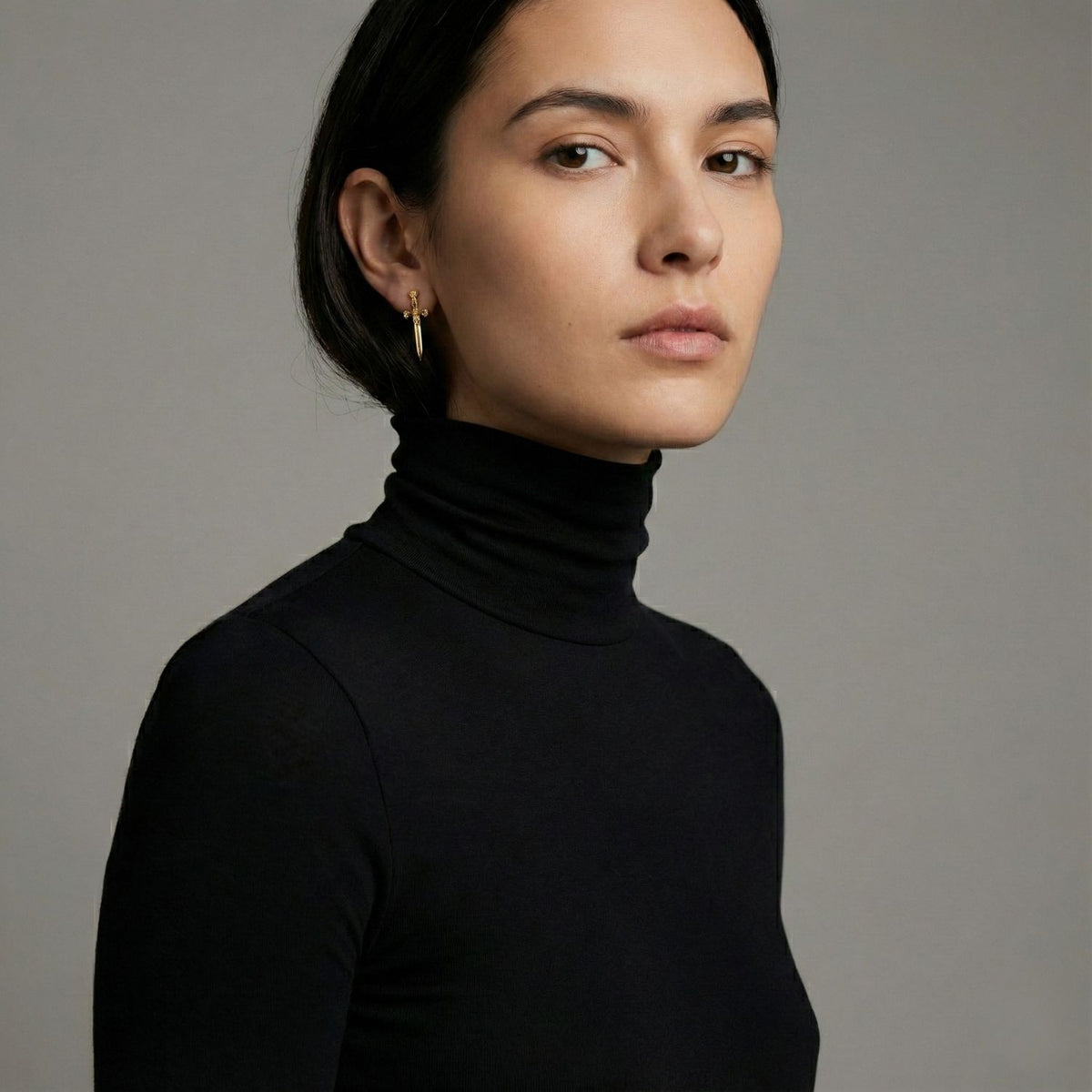 A woman with straight dark hair tucked behind her ears wears a black turtleneck and JAREDJAMIN Jewelry Online’s 18K Gold Lady Macbeth Stud Earrings, looking slightly to the side against a plain light gray background.