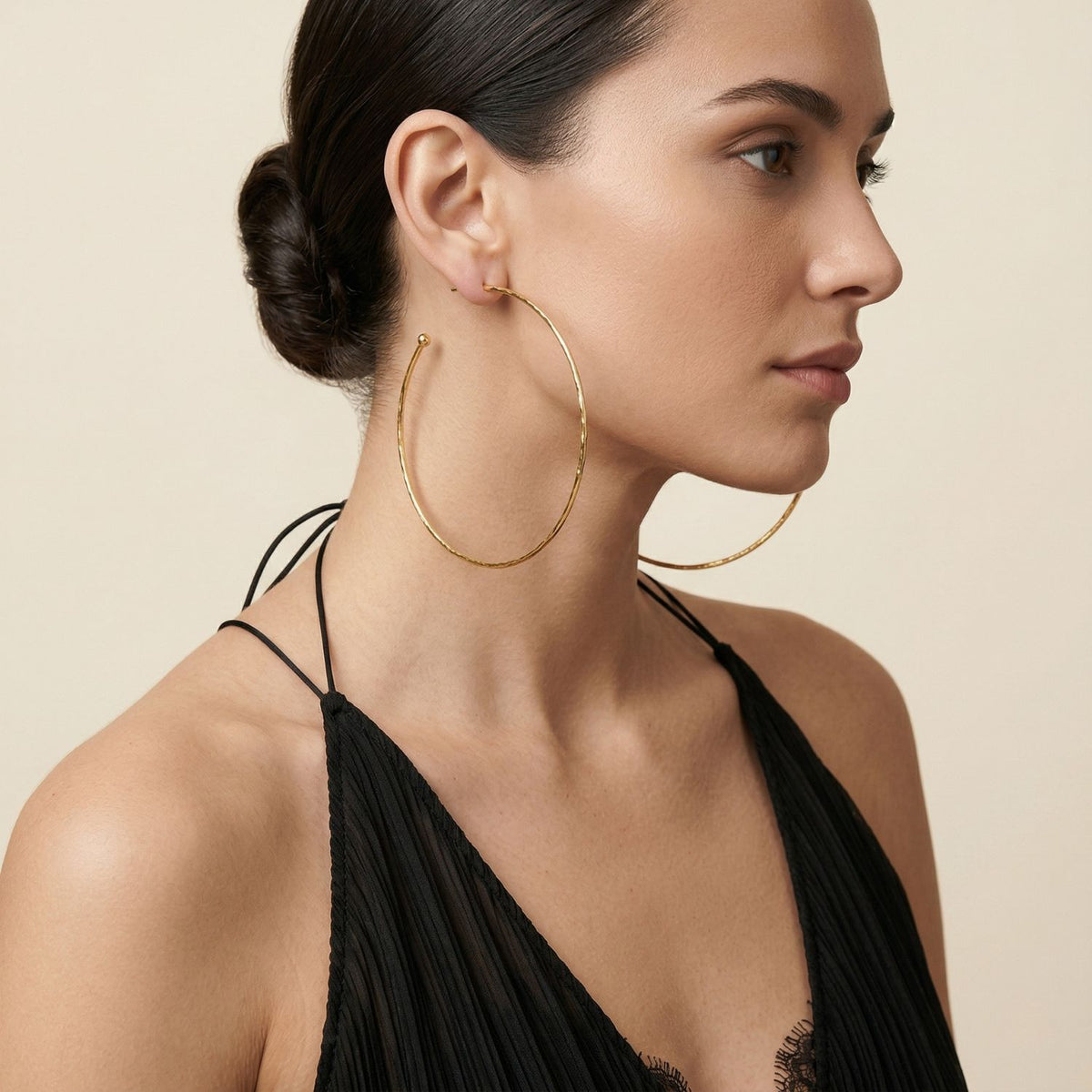 A woman with dark hair in a low bun wears a black halter top and our Gold Large Hooptastic Hoop Post Earrings—hand-assembled in West Hollywood using small-batch, eco-conscious methods. Style Note: punctuates both minimal tailoring and evening silks.