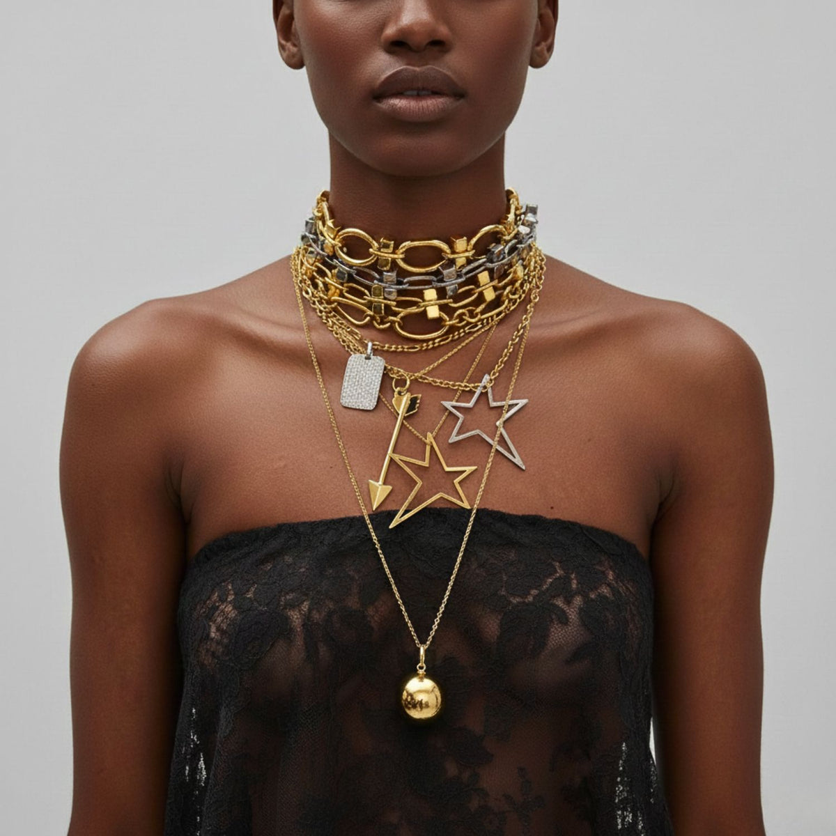 Un woman en un black lace, strapless top layers chunky oro y plata chains, including JAREDJAMIN Joyería Online’s plata Large Shooting Star colgante dije. Her partially visible face stands out against the plain light gray background.