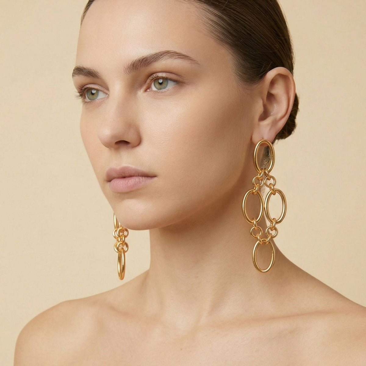 Un light-skinned, light-eyed woman avec slicked-back brown hair dans un low bun wears large or Ophelia pendante Post boucles d'oreilles par JAREDJAMIN Bijoux en Ligne. She gazes slightly to the side against a neutral beige background, her shoulders bare.
