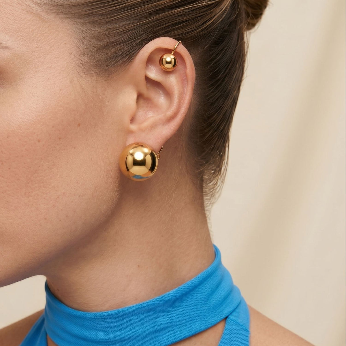 Side profile view of a model wearing the Gold Orbita Ear Cuff positioned along the upper helix, demonstrating its non-pierced floating sphere illusion and bright metallic reflection paired with a vibrant blue collar top.