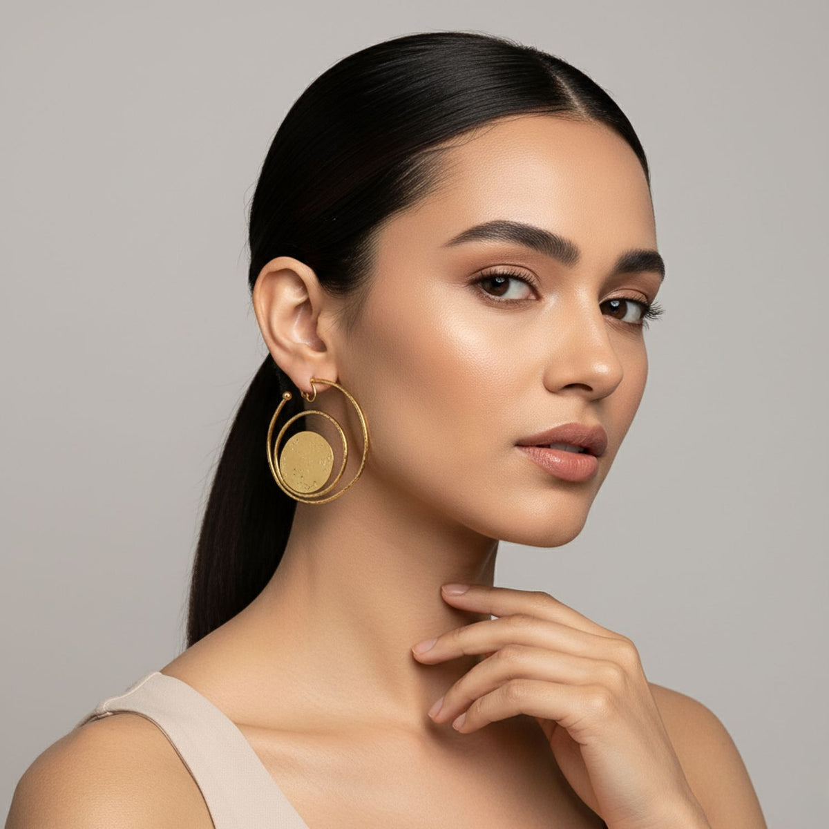 A woman with smooth, straight dark hair in a low ponytail wears JAREDJAMIN Jewelry Online’s Gold Peacock Clip On Hoop Earrings and a sleeveless beige top. Her makeup is natural with defined brows, subtle eyeshadow, and nude lips.