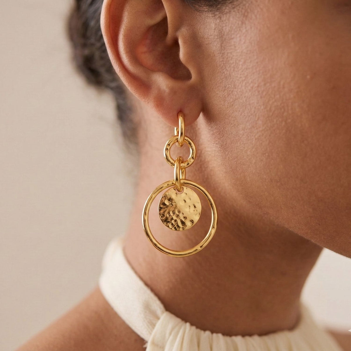 A close-up shows a person with medium skin and dark, pulled-back hair wearing JAREDJAMIN Jewelry Online’s Gold Peacock Preen Post Earrings—a handcrafted pair with a hammered metal disc and large gold hoop—over a light halter-neck top against a neutral background.