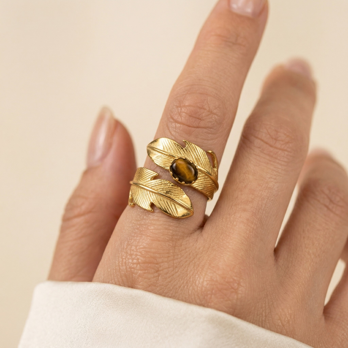 A medium-toned hand in an off-white sleeve punctuates the scene, adorned with our Gold Plume Adjustable Ring—hand-assembled in our West Hollywood studio. Sculpted leaves cradle a brown gemstone. Small-batch, eco-conscious. Style Note: Equally at home with denim or silk.