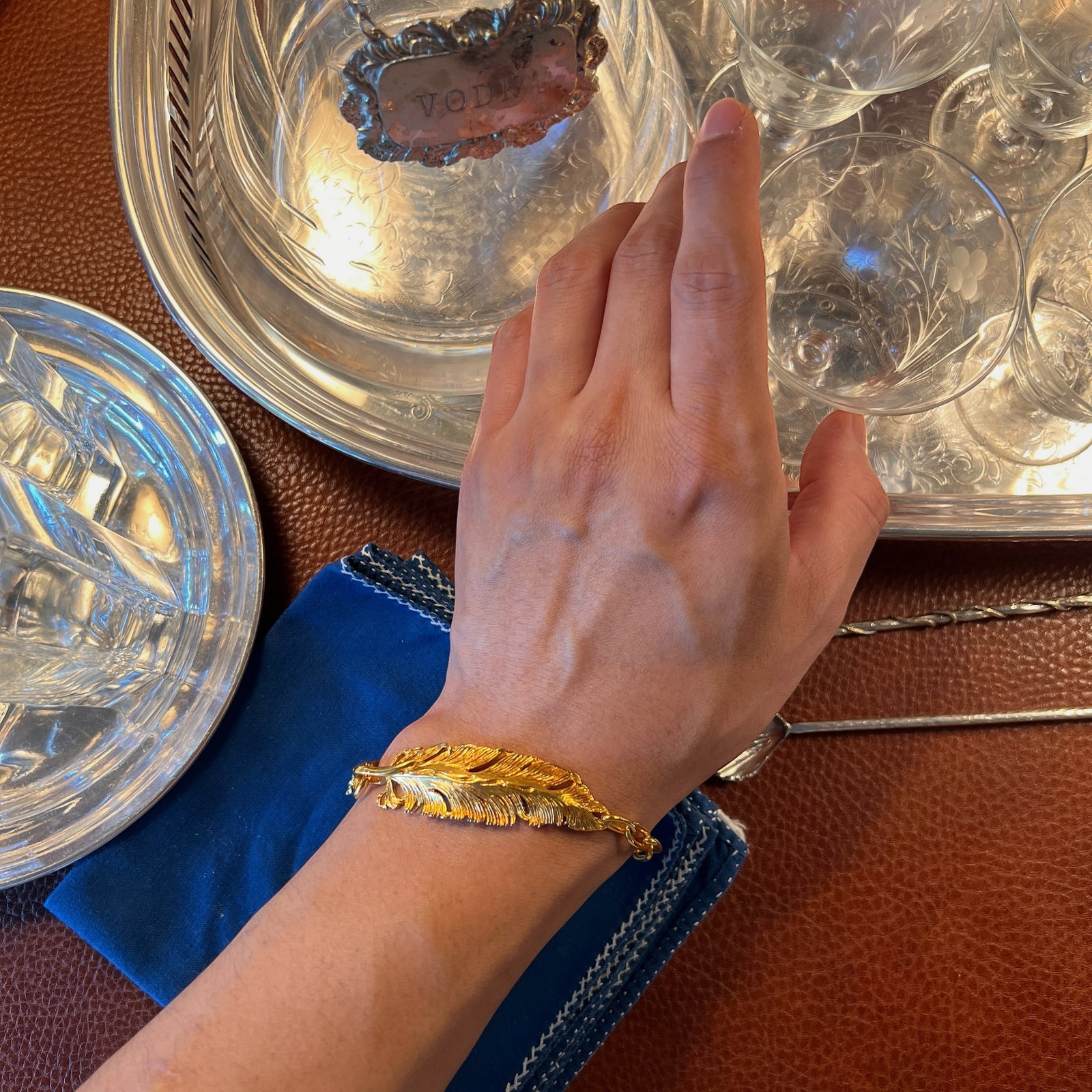 The Gold Feather Plume Bracelet by JAREDJAMIN Jewelry Online features an oversized, detailed feather on a chunky chain with a small rectangular tag for a personalized touch, exuding whimsical elegance against a plain white background.