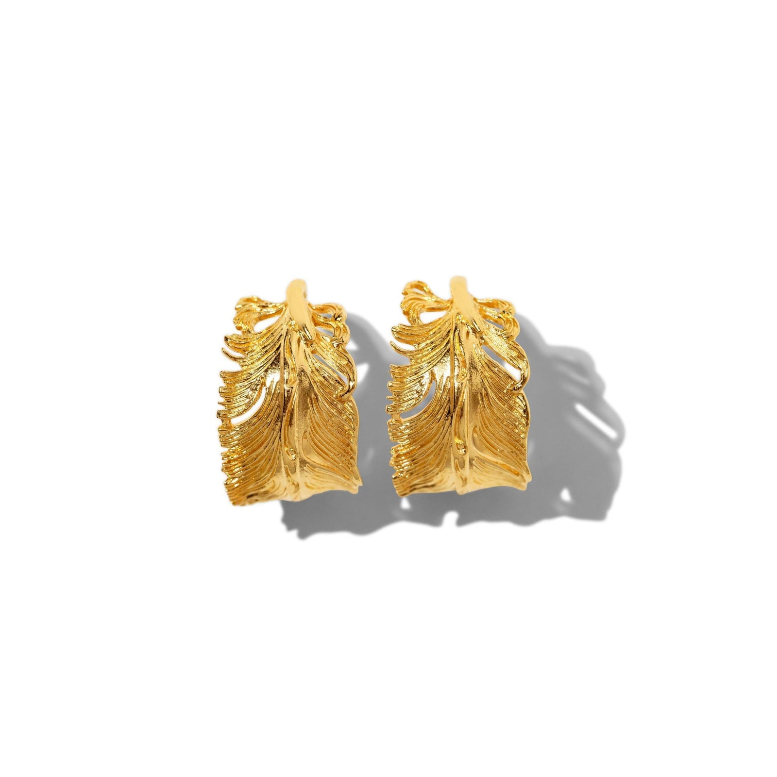 The Gold Plume Hoop Post Earrings by JAREDJAMIN Jewelry Online feature detailed, textured leaf designs with intricate veins and edges, resembling golden feathers. Shown upright on a white background, they cast soft right-facing shadows.