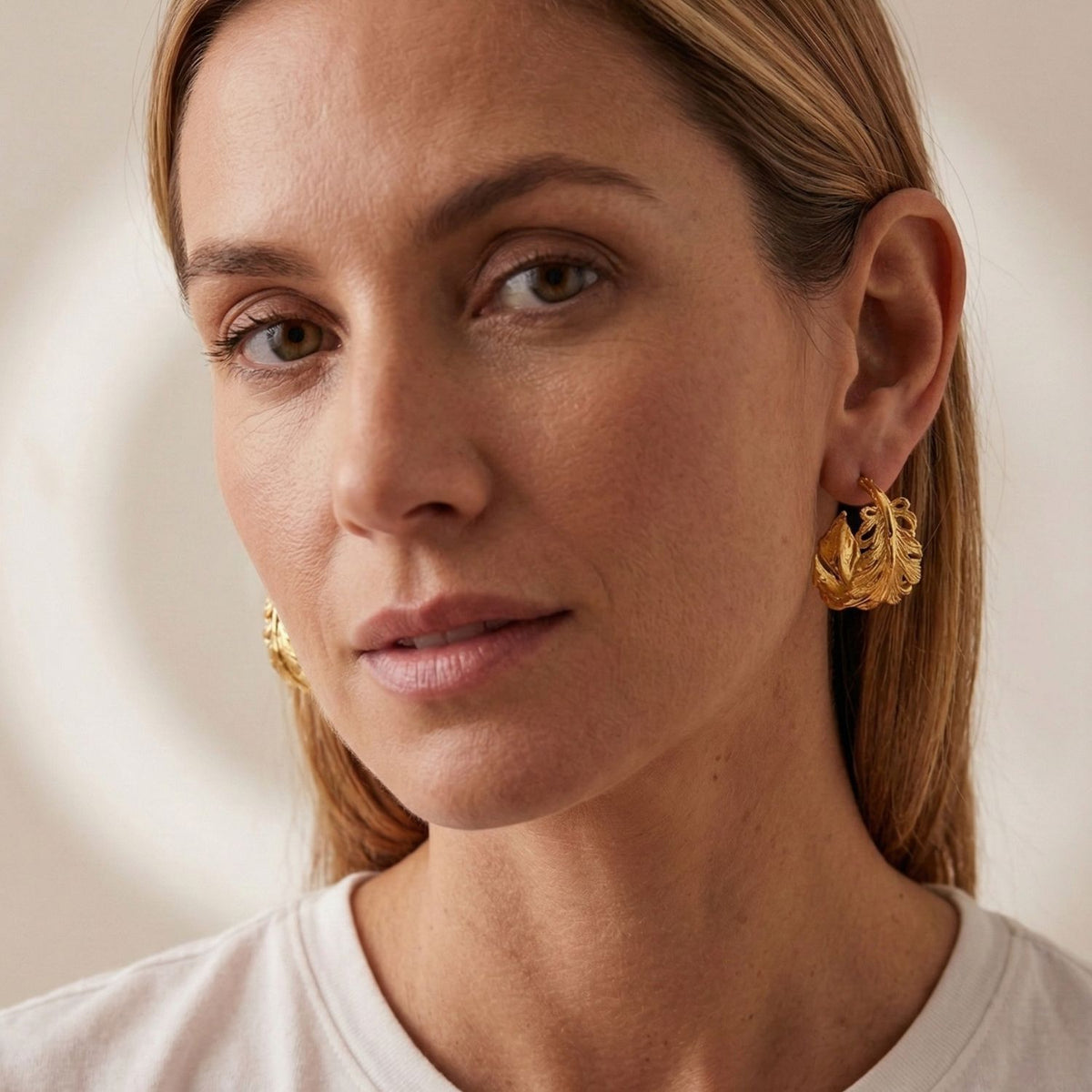 JAREDJAMIN Jewelry Online's Gold Plume Hoop Post Earrings, featuring large 18K gold-plated leaf shapes, are worn with a white top. The scene is set against a soft background for a calm, minimalist effect.