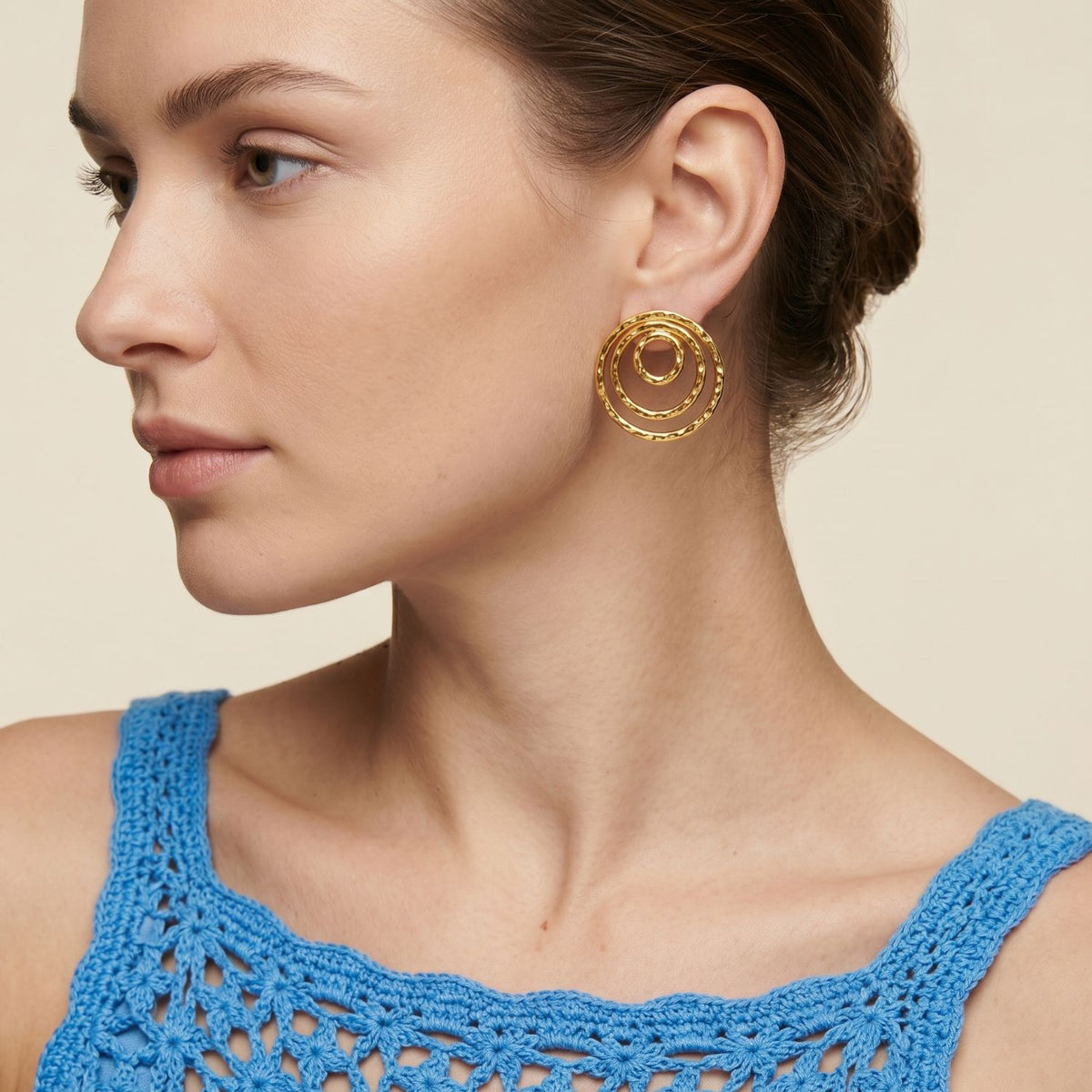 A woman with fair skin and a low brown bun punctuates a blue crochet top with our Gold Ripple Textured Clip On Earrings—hand-assembled in our West Hollywood studio, rendered in eco-minded small batches. Style Note: transitions effortlessly from tailored blazers to silk tanks.