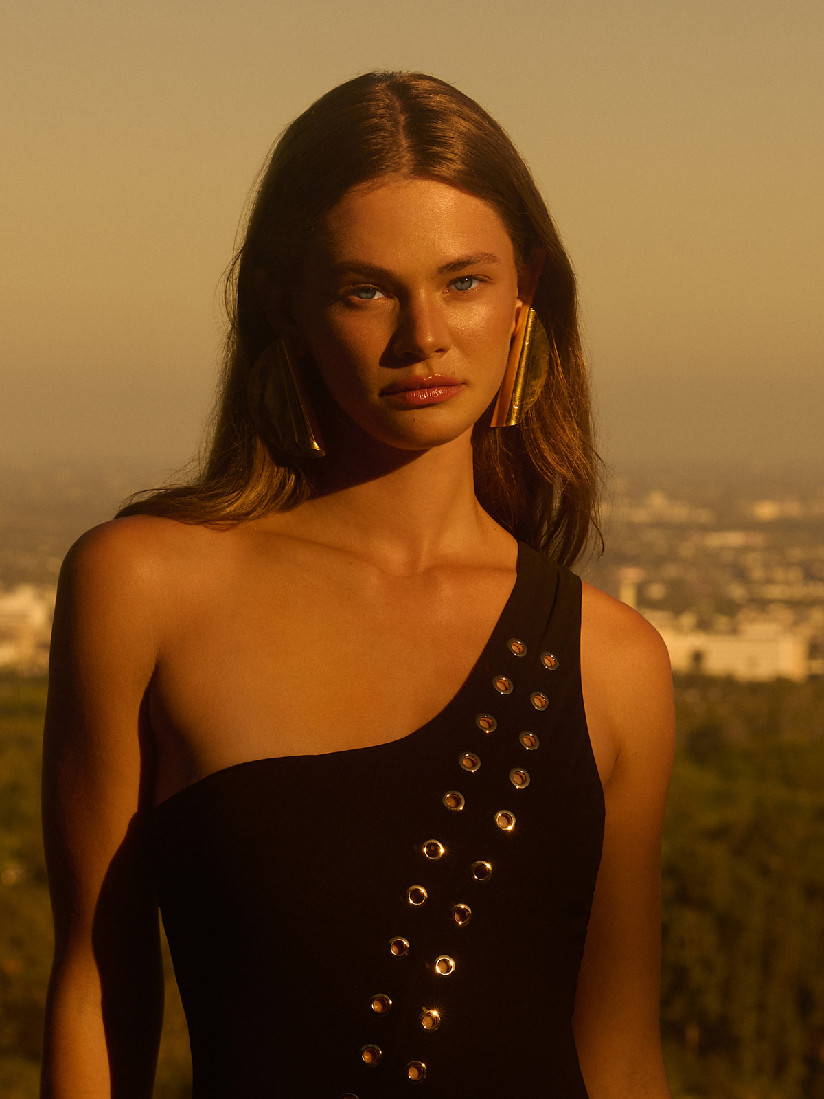 A woman stands outdoors at sunset in a black one-shoulder dress with metallic accents, featuring long brown hair and large Scroll Earrings by JAREDJAMIN Jewelry Online. The backdrop showcases a blurred cityscape beneath a warm, golden sky.