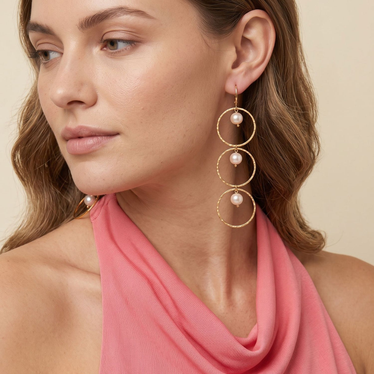 Close-up of a woman wearing one gold dangle earring with stacked hammered hoops and pearl accents; pink coral top.