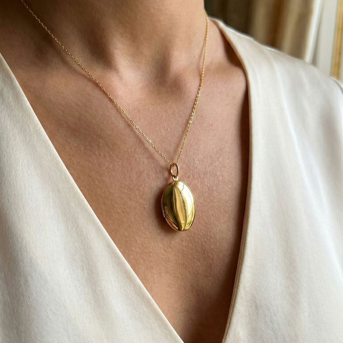 Un primer plano shows a person wearing the JAREDJAMIN Joyería Online oro Smooth collar colgante Locket, an elegante 18K bañado en oro locket sobre un delicado cadena, centered over a cream V-neck blouse against a softly blurred background.
