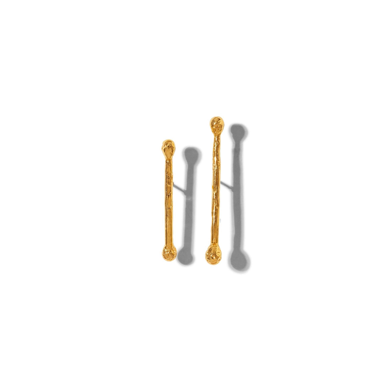 The Gold Stix Post Earrings by JAREDJAMIN Jewelry Online feature two textured, gold-colored metal sticks with rounded ends and 18K Gold posts, arranged vertically on white to showcase their unique, organic surfaces and soft gray shadows.