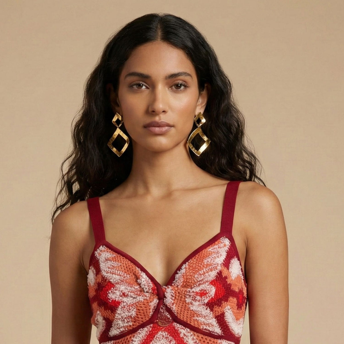 A woman with long wavy dark hair wears large Gold Toscana Diamond Drop Post Earrings by JAREDJAMIN Jewelry Online. She has a calm expression, is dressed in a sleeveless red and pink patterned top, and stands against a plain beige background.