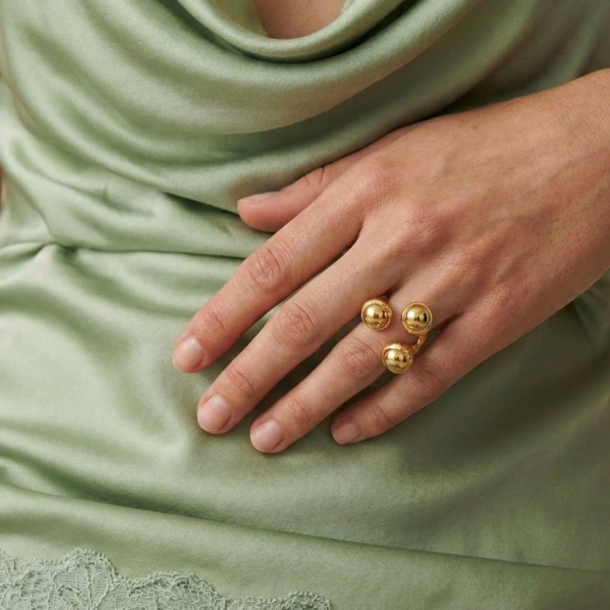 Overhead view of a model's hand wearing the Gold Trinity Acorn Statement Ring, showcasing its dimensional botanical scale and warm metallic glow styled with a sage green satin dress.