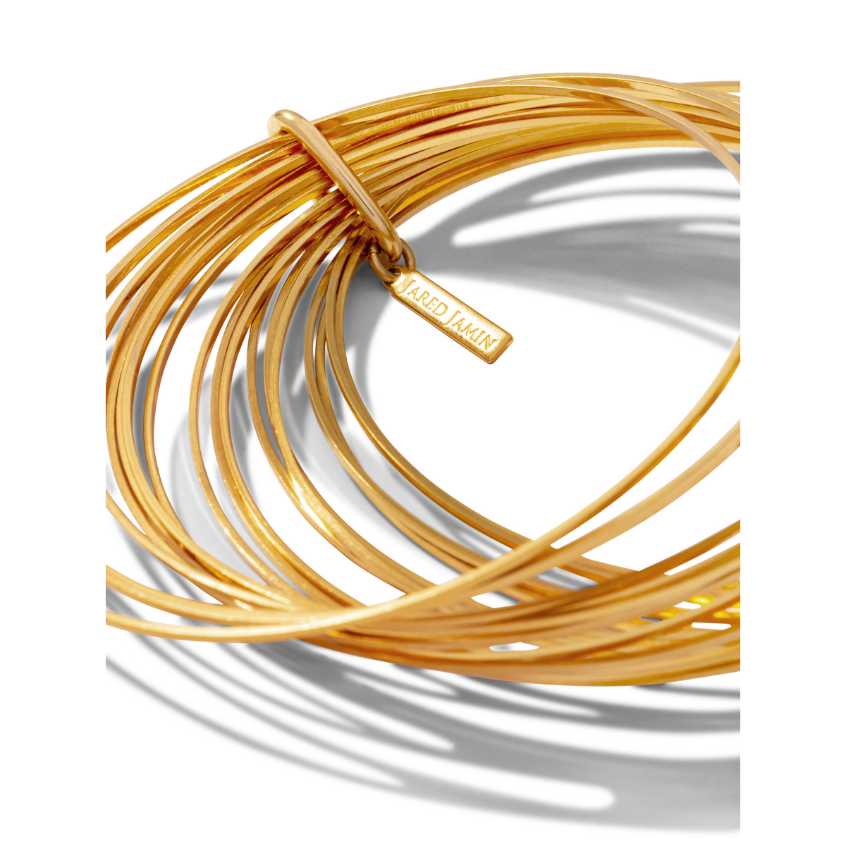 Les or Twenty Something jonc Stack par JAREDJAMIN Bijoux en Ligne features thin, interlocking 18K plaqué or joncs. One jonc has a “JARED JAMIN” tag, arranged dans un circular stack et casting intricate shadows sur un fond blanc.
