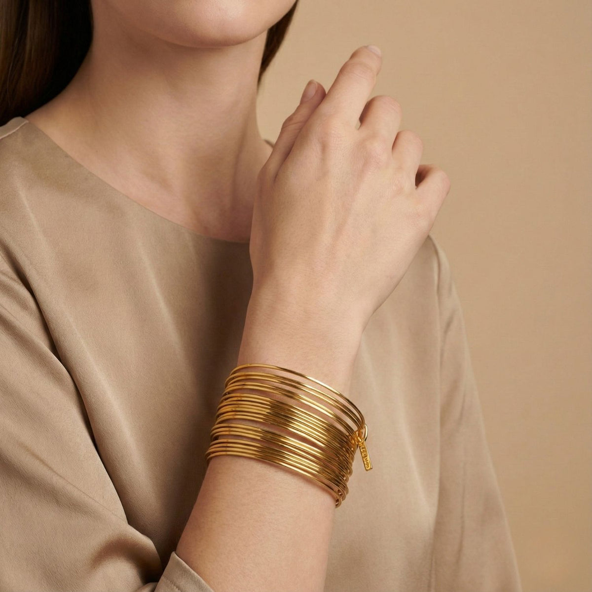 A woman in a beige top poses with her hand near her face, highlighting the Gold Twenty Something Bangle Stack by JAREDJAMIN Jewelry Online. The neutral background complements the elegant 18K gold-plated bangles. Her face is partially visible.