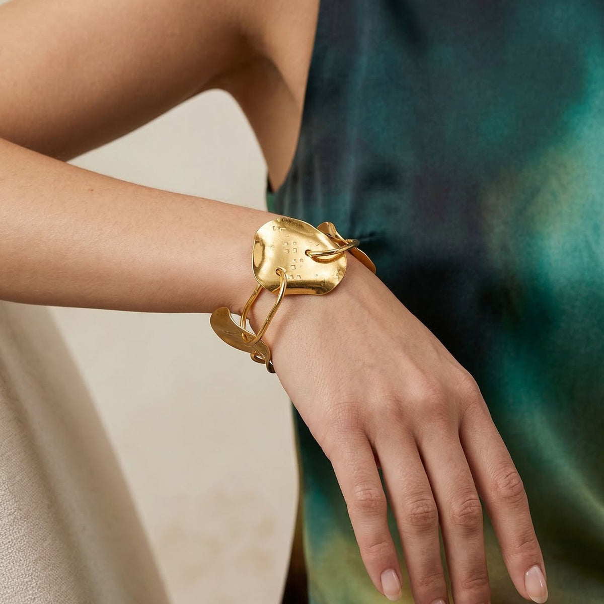 The Gold Viviane Link Bracelet, a handcrafted statement piece with engraved text and two abstract, irregular gold links, is displayed on a neutral surface, modeled by someone in a sleeveless green-patterned dress.