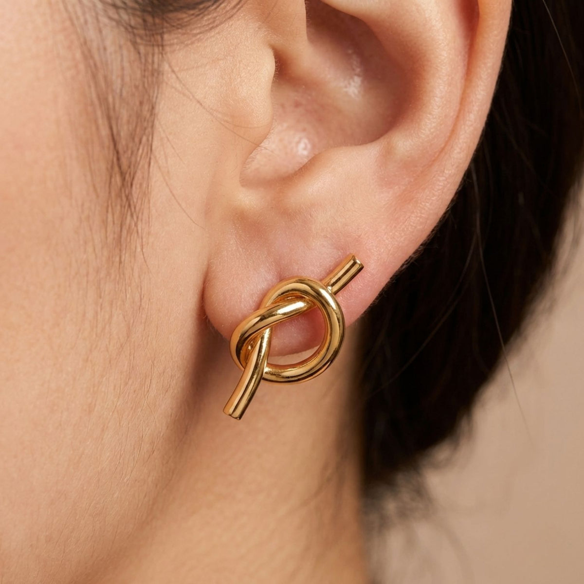 Close-up of a person's pierced ear wearing JAREDJAMIN Jewelry Online's Gold Why Knot Stud Post Earrings. The 18K gold-plated knot design features intersecting loops for a modern touch, set against a neutral background with visible dark hair.