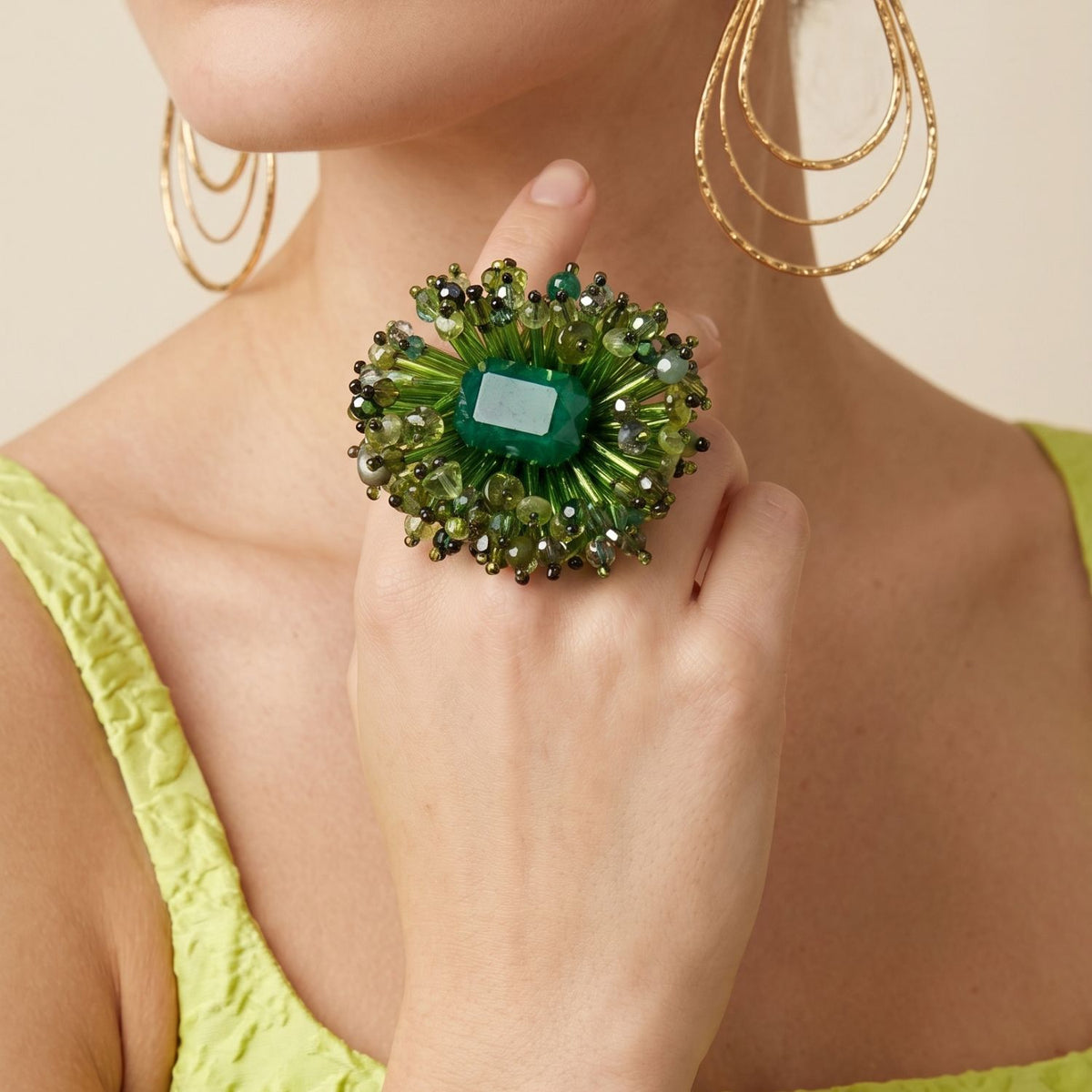 Close-up portrait of a female model wearing a bright green top and gold earrings, styling the oversized Ivy Green Beaded Burst Ring on her hand to demonstrate its dramatic, sunburst-like scale and vibrant color palette.