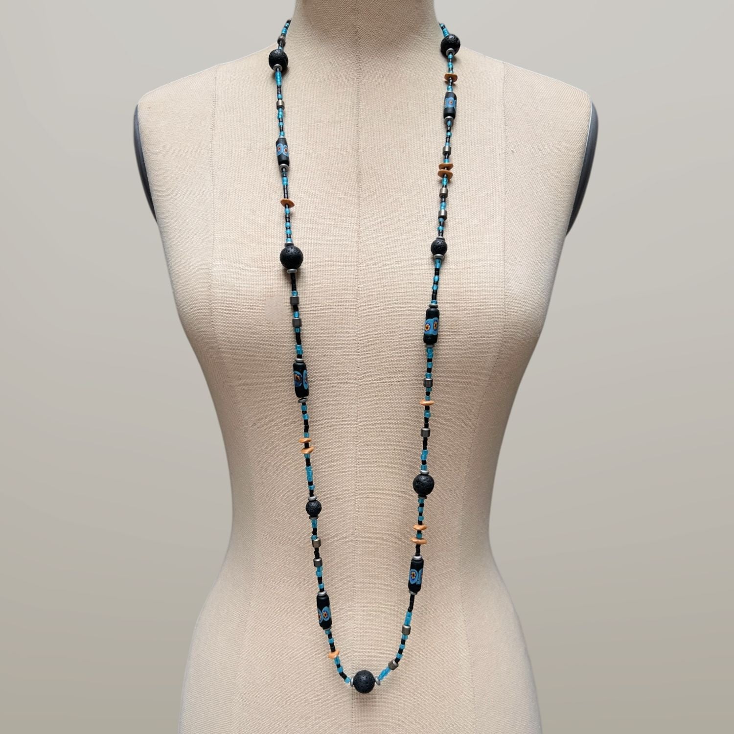 The African Voyage Long Lava Necklace by JAREDJAMIN Jewelry Online features three vintage strands with black lava, hematite, blue hand-painted African glass beads with floral patterns, sky-blue and black seed beads, tan discs, and a silver clasp.