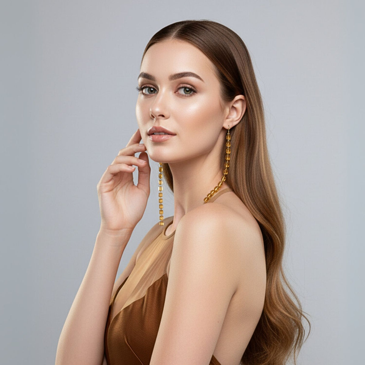 Un woman con long, wavy brown hair poses in profile against a light gray background, wearing a sleeveless brown dress y JAREDJAMIN Joyería Online’s Amber Building Block colgante pendientes, her makeup natural as she touches her face y looks confidently at the camera.