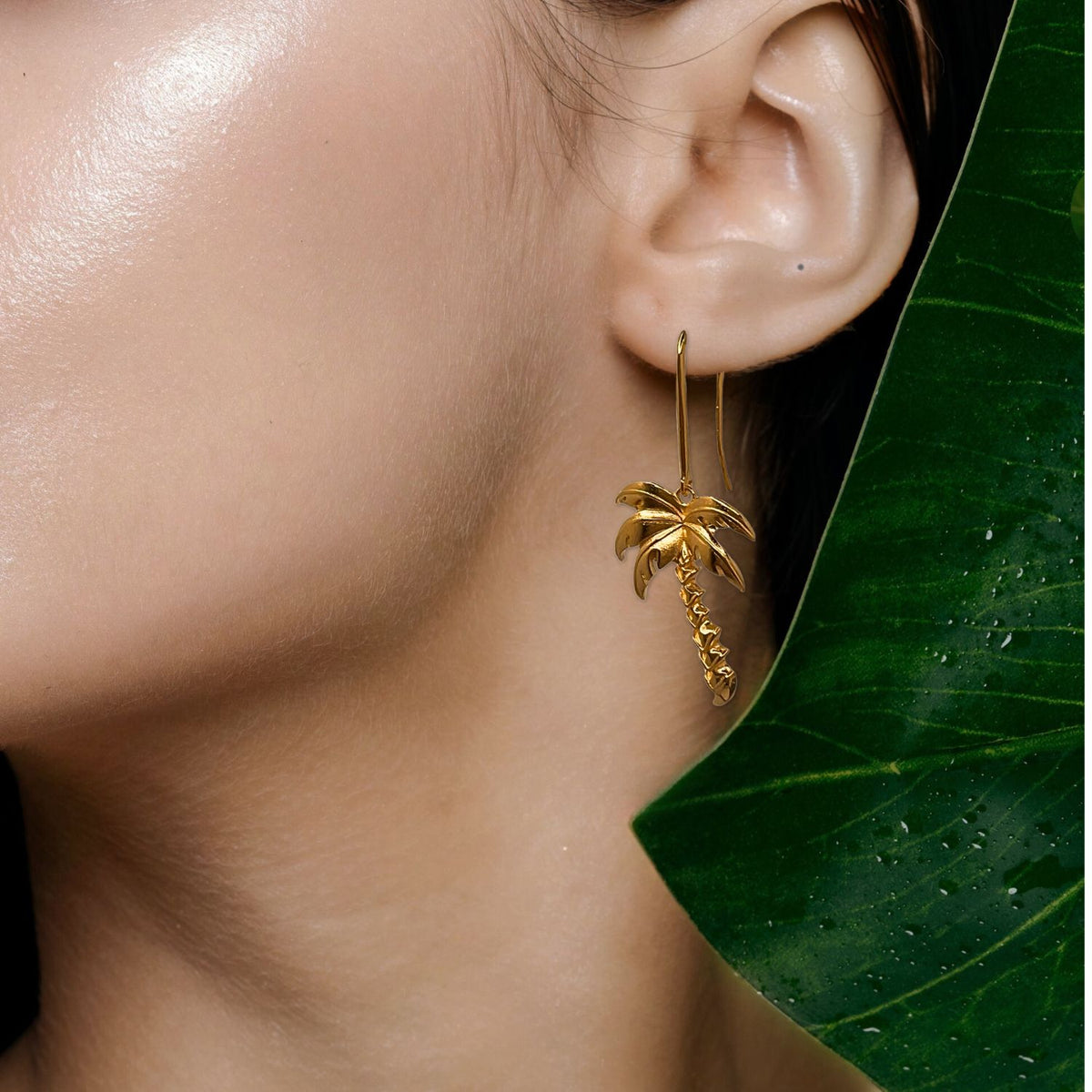 A close-up of a woman&#39;s ear showcases JAREDJAMIN Jewelry Online&#39;s Gold Angeleno Palm Hook Earrings. Her hair is pulled back, and a shiny green leaf with water droplets partially frames the scene.