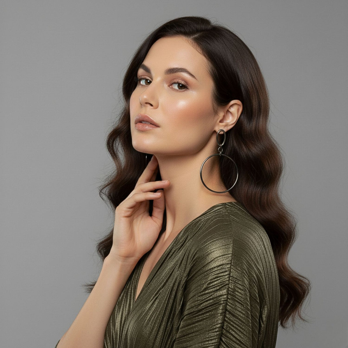 Un woman con long, wavy brown hair poses against a gray background wearing a shimmery olive-green blouse y JAREDJAMIN Joyería Online’s Antique Ophelia colgante aro Post pendientes, her hand gently touching her neck y her makeup soft y glowing.