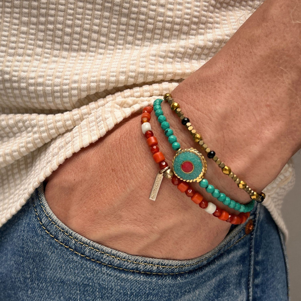 A hand in a blue jean pocket showcases the JAREDJAMIN Jewelry Online Aqua Evil Eye Bracelet Set, a vibrant trio featuring colorful beads with turquoise and gemstone accents, paired with a textured cream shirt.