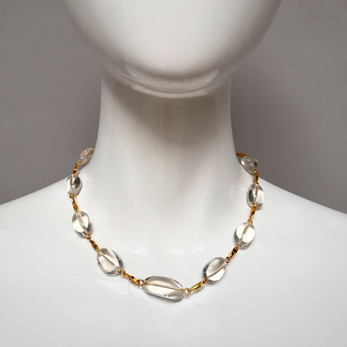 The Arctic Gold Bar-Link Necklace by JAREDJAMIN Jewelry Online adorns a mannequin bust, featuring clear oval crystal quartz beads with small gold connectors. The beads subtly reflect light, creating a simple elegance, with an 18K gold plated toggle clasp against a neutral gray background.