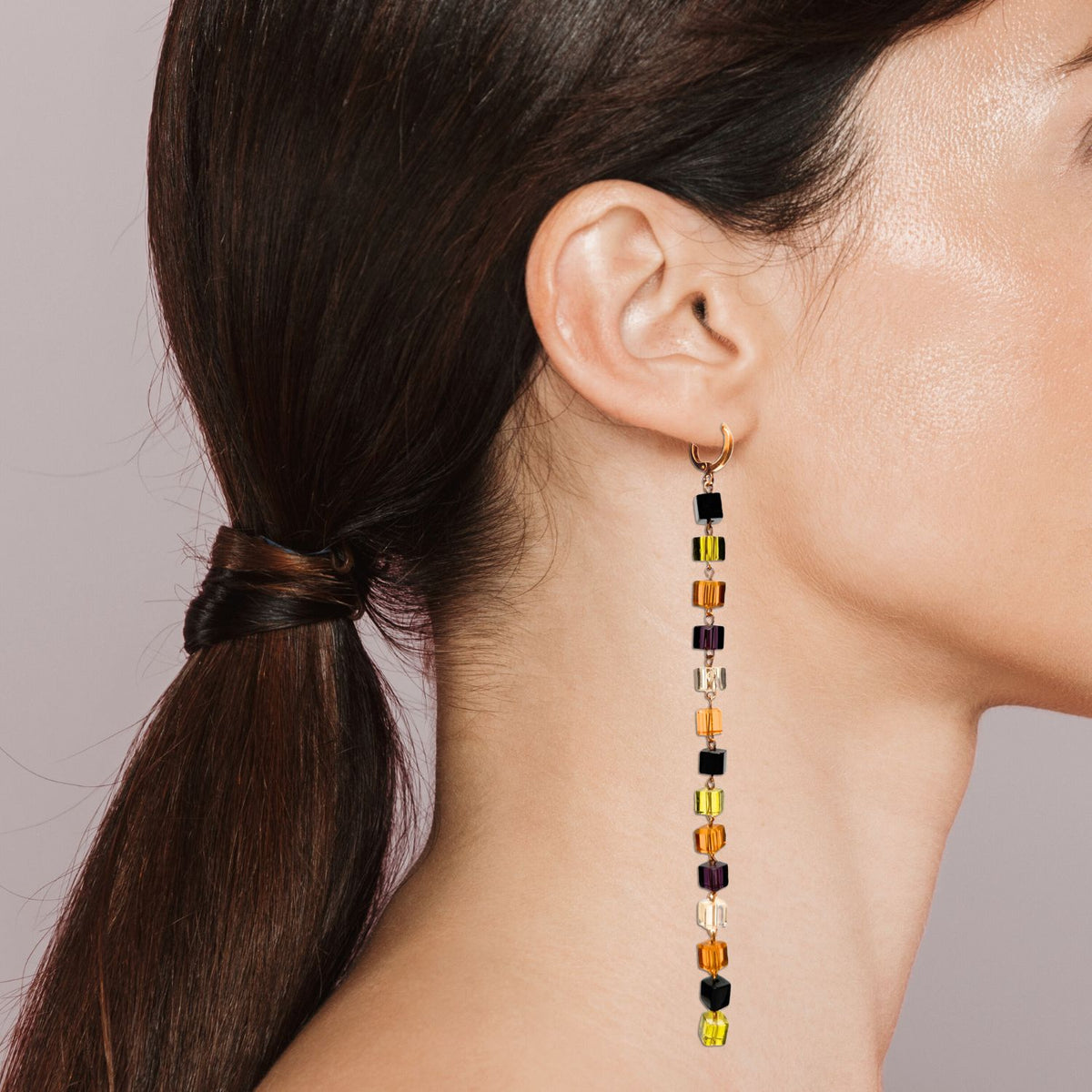 A woman with straight brown hair in a low ponytail showcases JAREDJAMIN Jewelry Online’s Auburn Building Block Drop Earrings, featuring multicolored beads, in profile against a plain light background.