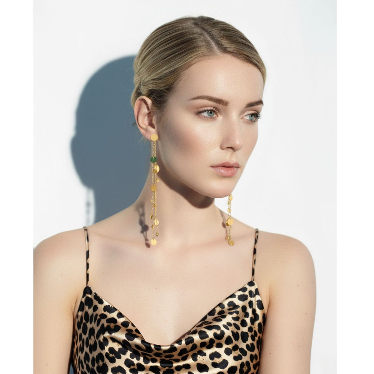A fair-skinned, blonde woman with a low bun wears JAREDJAMIN Jewelry Online's Babylonica Dangle Post Earrings and a leopard print satin camisole. She faces right with a neutral expression, bright light casting a soft shadow on the white background.