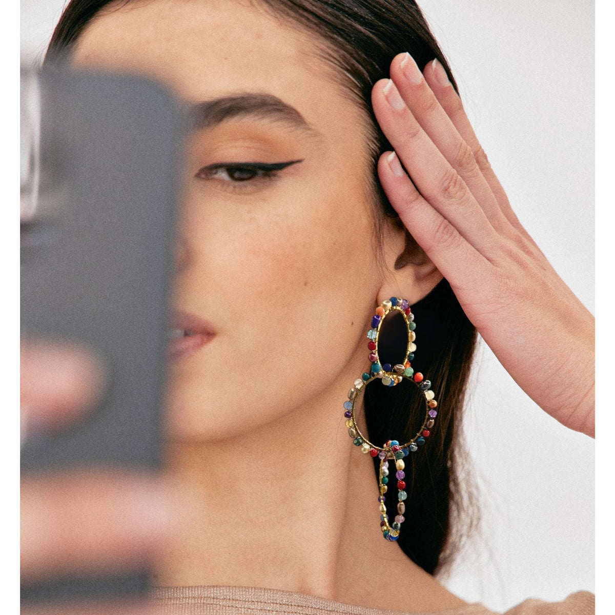 A woman with straight dark hair takes a selfie, wearing bold winged eyeliner and JAREDJAMIN Jewelry Online’s Begemmed Lindy 18K Gold Plated Earrings—statement pieces of rings adorned with multicolored beads, highlighted as she tucks her hair behind her ear.