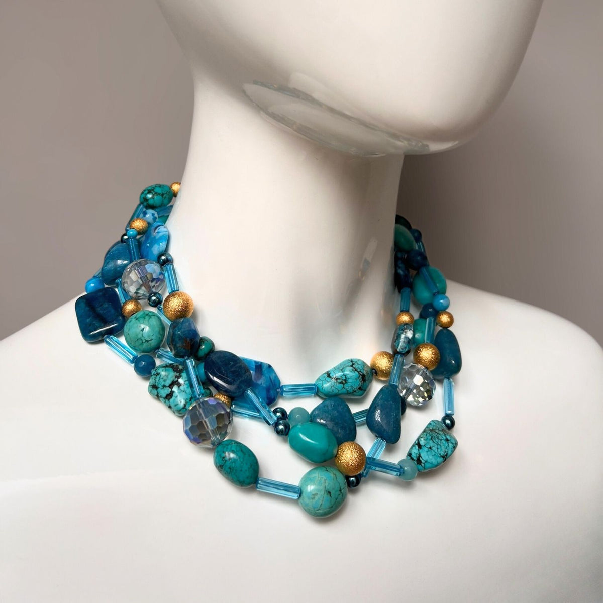 The Blue Reef Necklace by JAREDJAMIN Jewelry Online is a vibrant piece featuring turquoise stones, glass beads, and 18K Stardust beads. Shiny gold accents enhance its layered design against a soft, neutral background that beautifully highlights its striking colors.