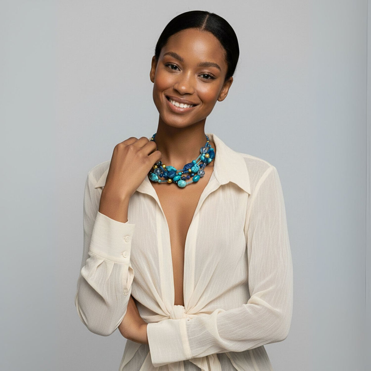 Un woman con dark hair pulled back smiles at the camera, wearing a sheer cream blouse y the Blue Reef collar de JAREDJAMIN Joyería Online. She gently touches the collar con her left hand against a light gray background.