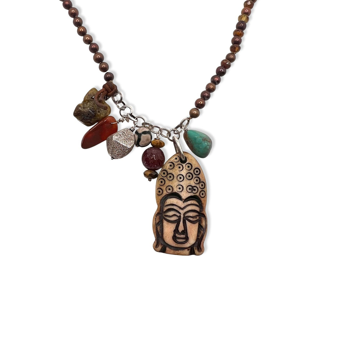 The Buddha Meanderings Beaded Necklace by JAREDJAMIN Jewelry Online features a carved wooden Buddha pendant with green, red, and brown jasper stones, a silver leaf charm, and a textured metal bead on a chain near the centerpiece.