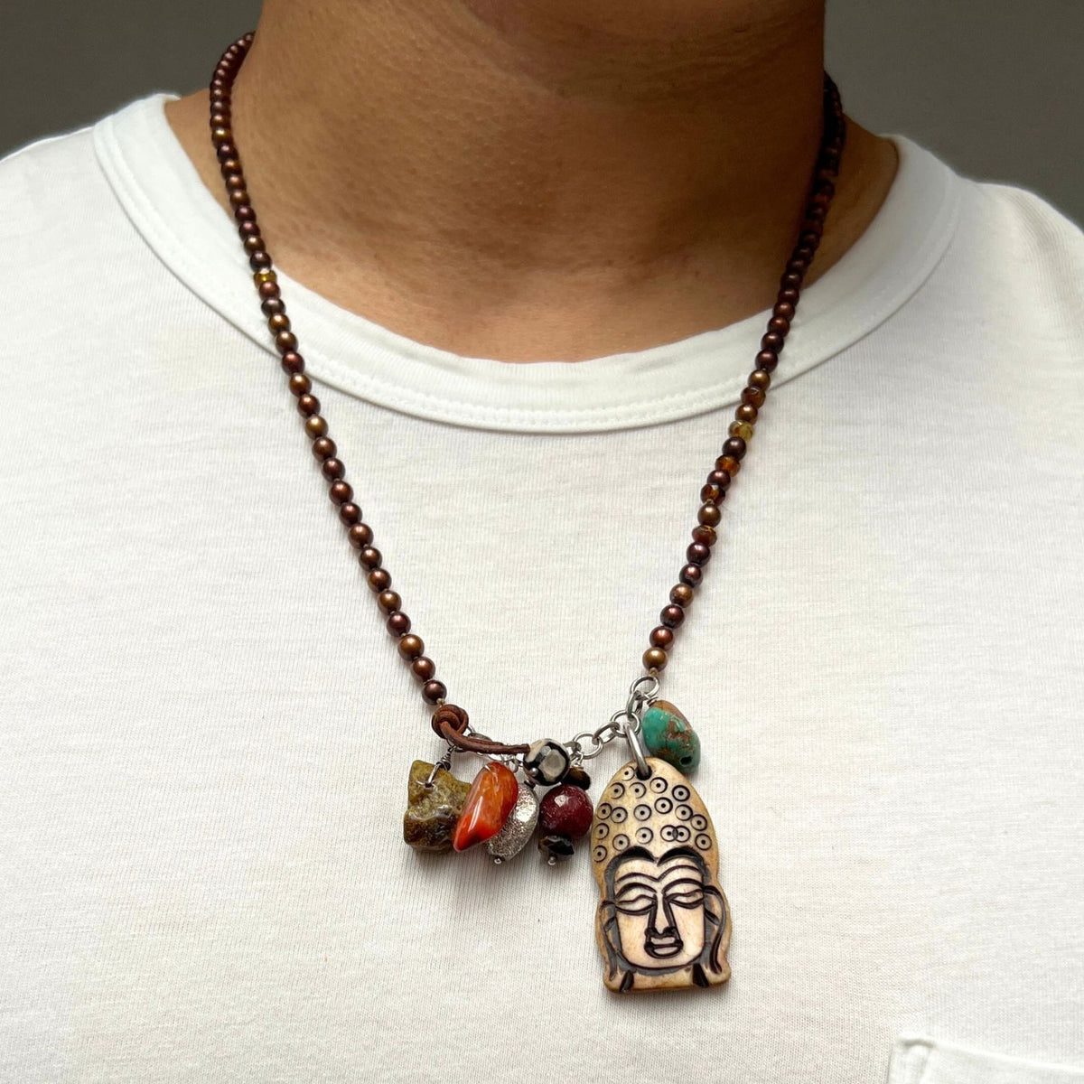 A person in a white shirt wears the Buddha Meanderings Beaded Necklace by JAREDJAMIN Jewelry Online, featuring a large Buddha head pendant with blue, orange, and brown jasper-like beads. The photo is closely cropped to the chest and neck.