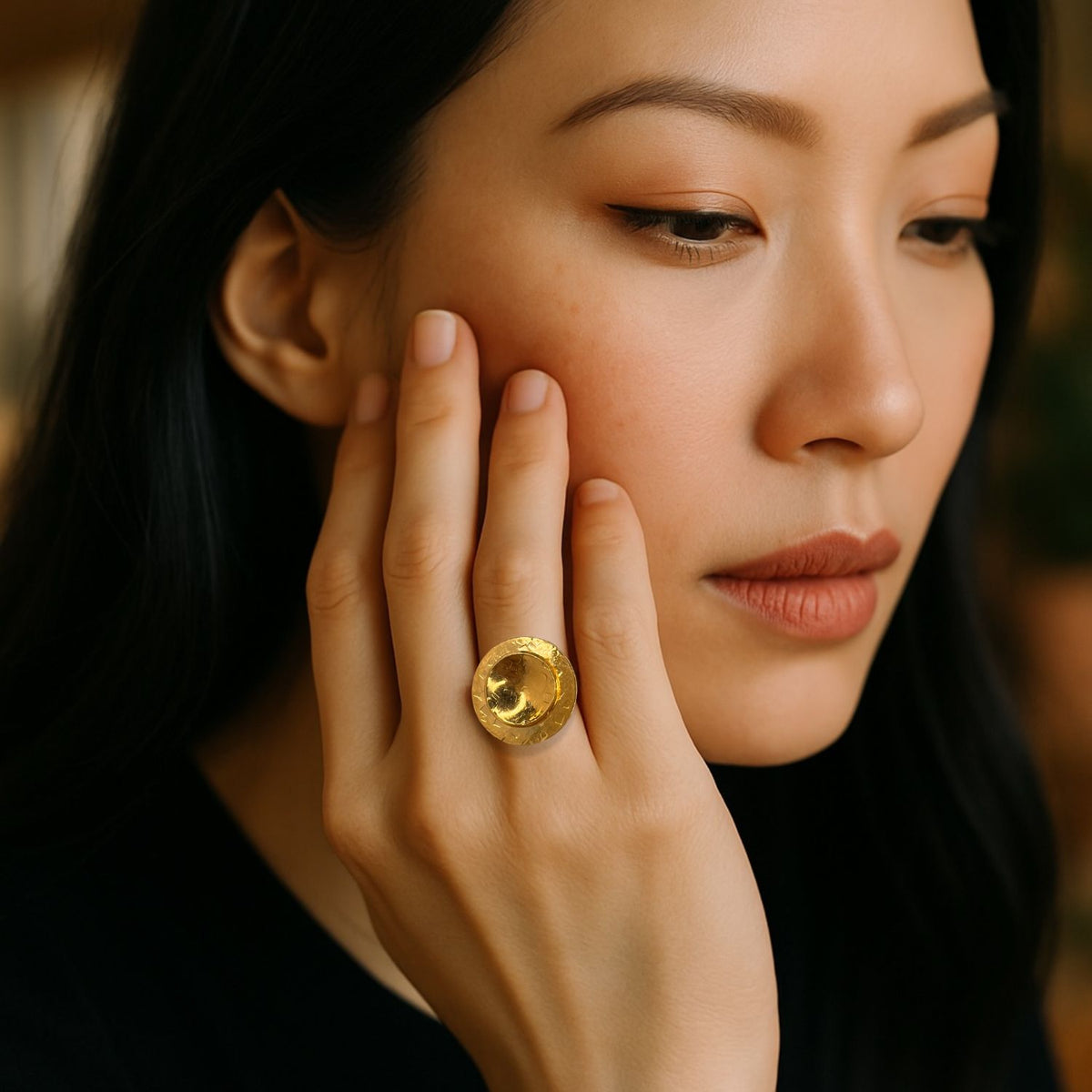 A woman with long black hair and a calm expression wears a black top and rests her hand on her face, elegantly showcasing the Buttercup Textured Adjustable Ring by JAREDJAMIN Jewelry Online. The 18K gold-plated design adds timeless style to her subtle look.