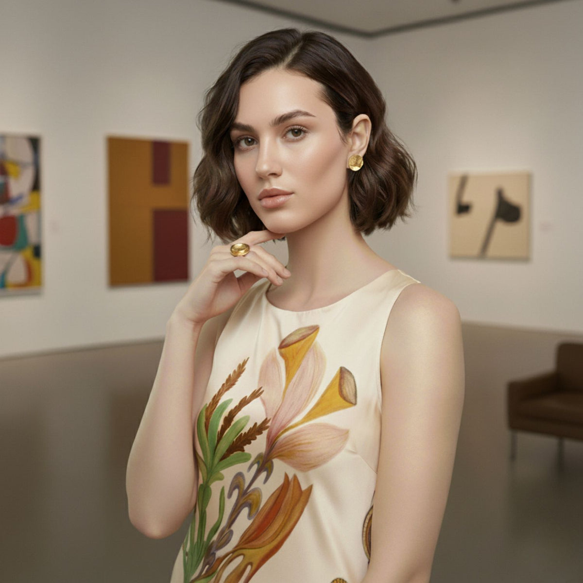 A woman with short brown hair and gold jewelry, including the JAREDJAMIN Jewelry Online Buttercup Textured Adjustable Ring, stands in an art gallery. She wears a sleeveless floral dress among abstract paintings and a brown couch.