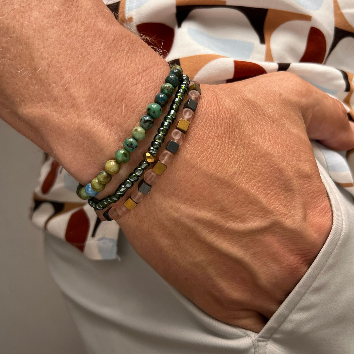 Un gros plan of a wrist wearing the Byzantine Rocks bracelet Set par JAREDJAMIN Bijoux en Ligne, avec beaded bracelets in green, or, et brown tones. Les hand rests in light beige pants avec a geometric patterned shirt.