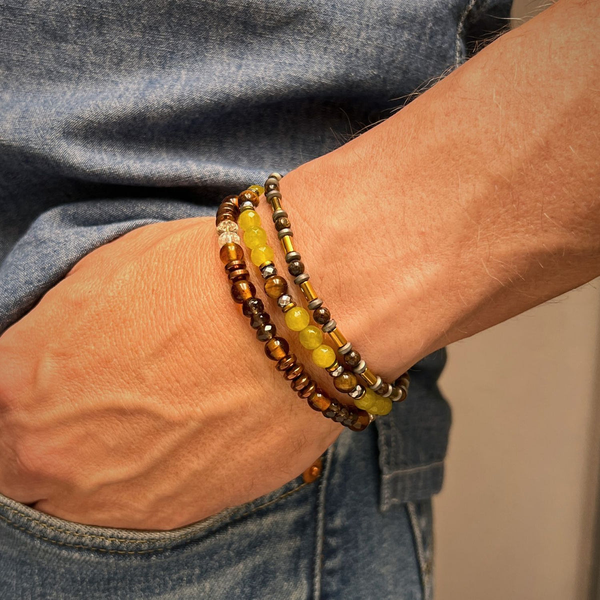 A close-up shows a hand in blue jeans pocket, wearing JAREDJAMIN Jewelry Online's Canyon Country Bracelet Set: three stretch bracelets in brown, yellow, and metallic tones including hematite, against a neutral background.