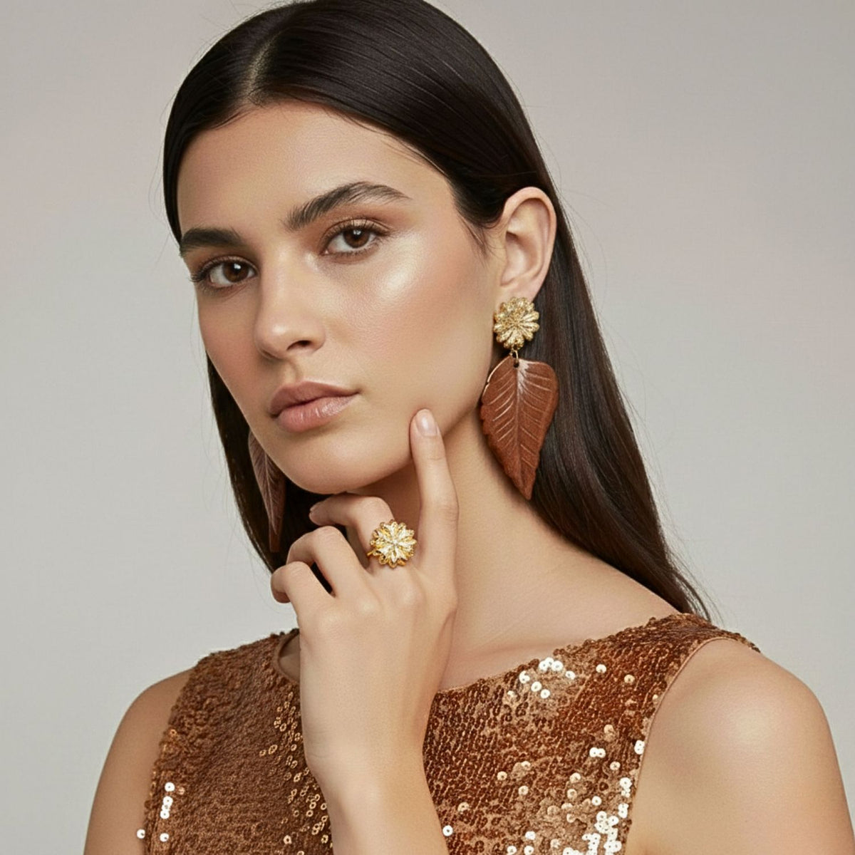 A womanとlong, straight dark hair poses confidently の中の sequined copper top, showcasing Chrysanthemum ゴールド Plated リング JAREDJAMINの ジュエリーオンラインとmatching イヤリング, her hand near her face highlighting 大胆な design.