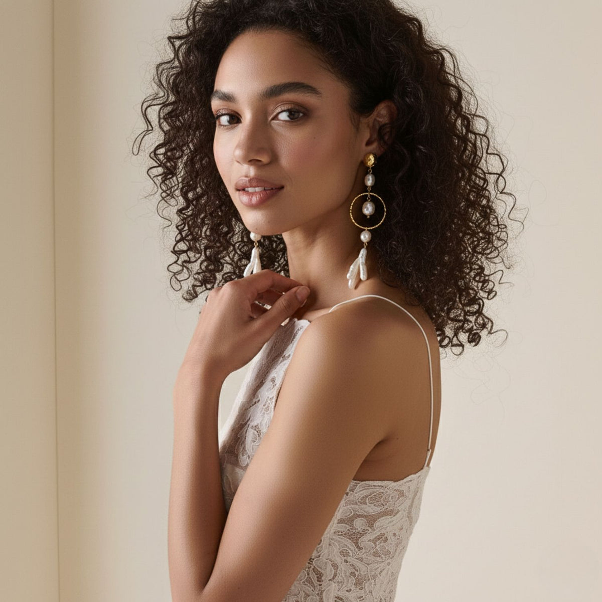 Un woman avec curly dark hair, wearing Ciara perle Dangle boucles d'oreilles par JAREDJAMIN Bijoux en Ligne et a white lace sleeveless dress, stands before a light background. She gazes softly at the camera, gently touching her collarbone.
