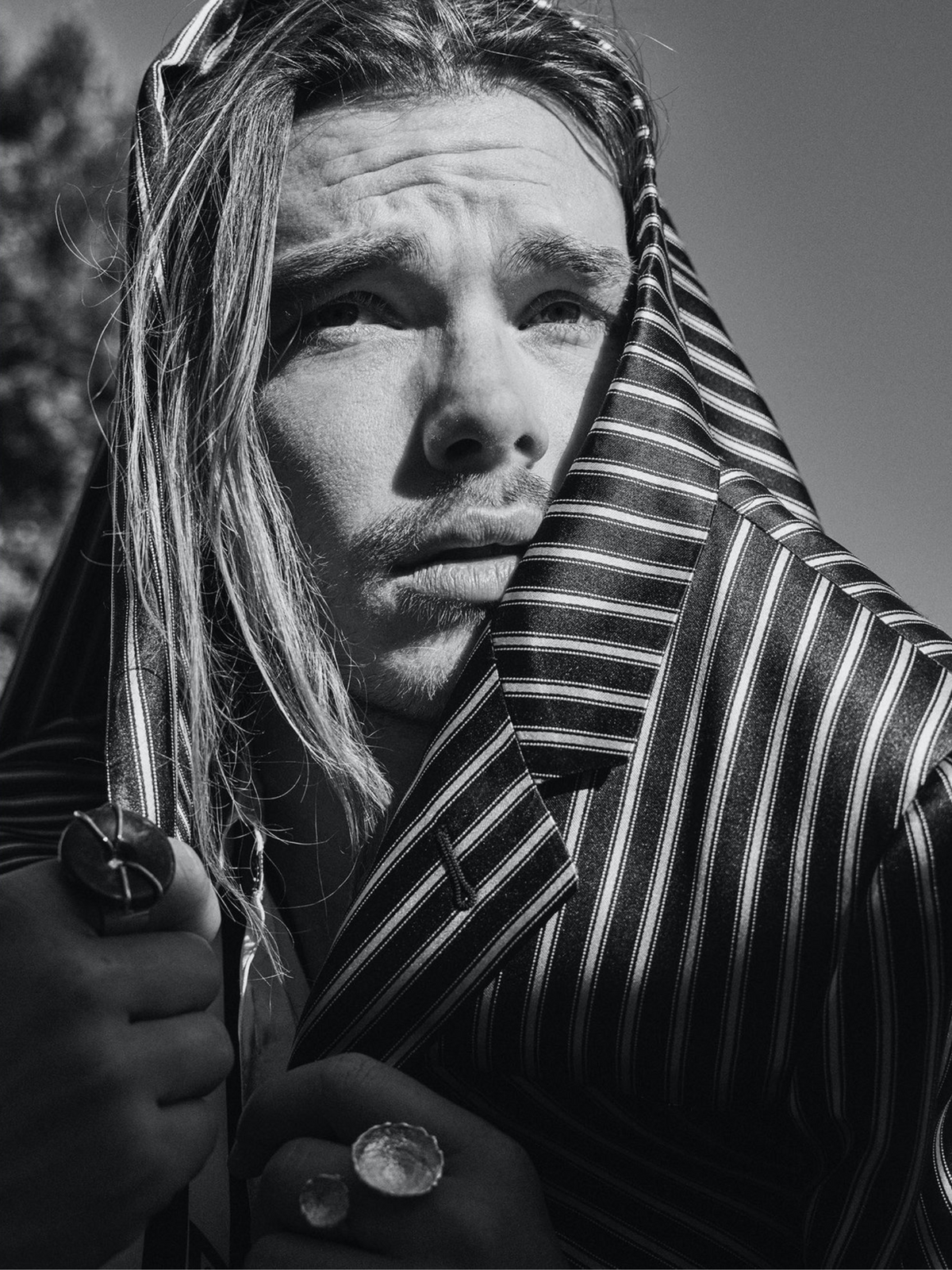 In a black and white portrait, a man with long hair draped in stripes gazes into the distance. He adorns several rings, including the Gold Cornelia Ring by JAREDJAMIN Jewelry Online, featuring Jasper stone. The image highlights contrast and texture with focused lighting on his contemplative face.